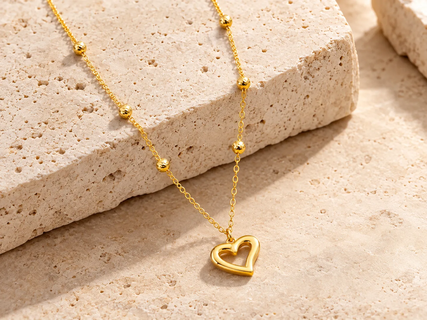 Fine gold chain with small round beads and an open heart pendant, resting on a stone ledge