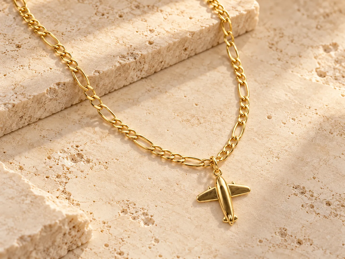 Gold necklace with an airplane-shaped pendant on a figaro-style chain, resting on a stone ledge
