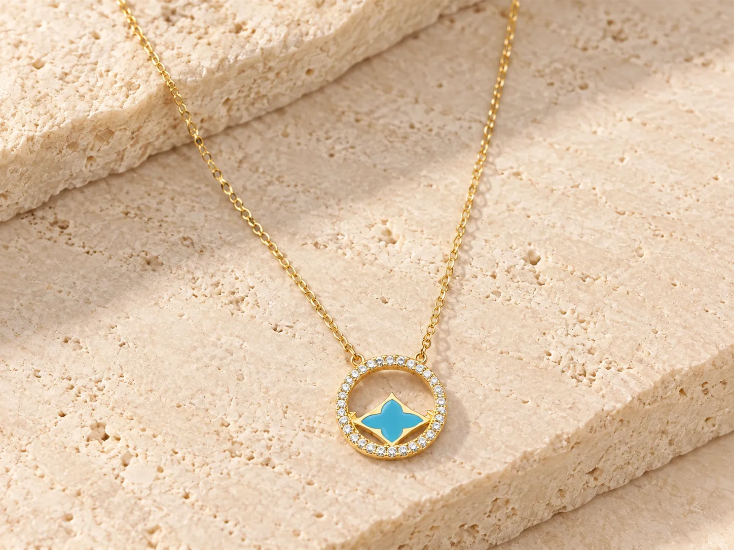 Gold necklace with a round halo pendant of small clear stones around a blue clover-shaped centre, on a warm stone surface