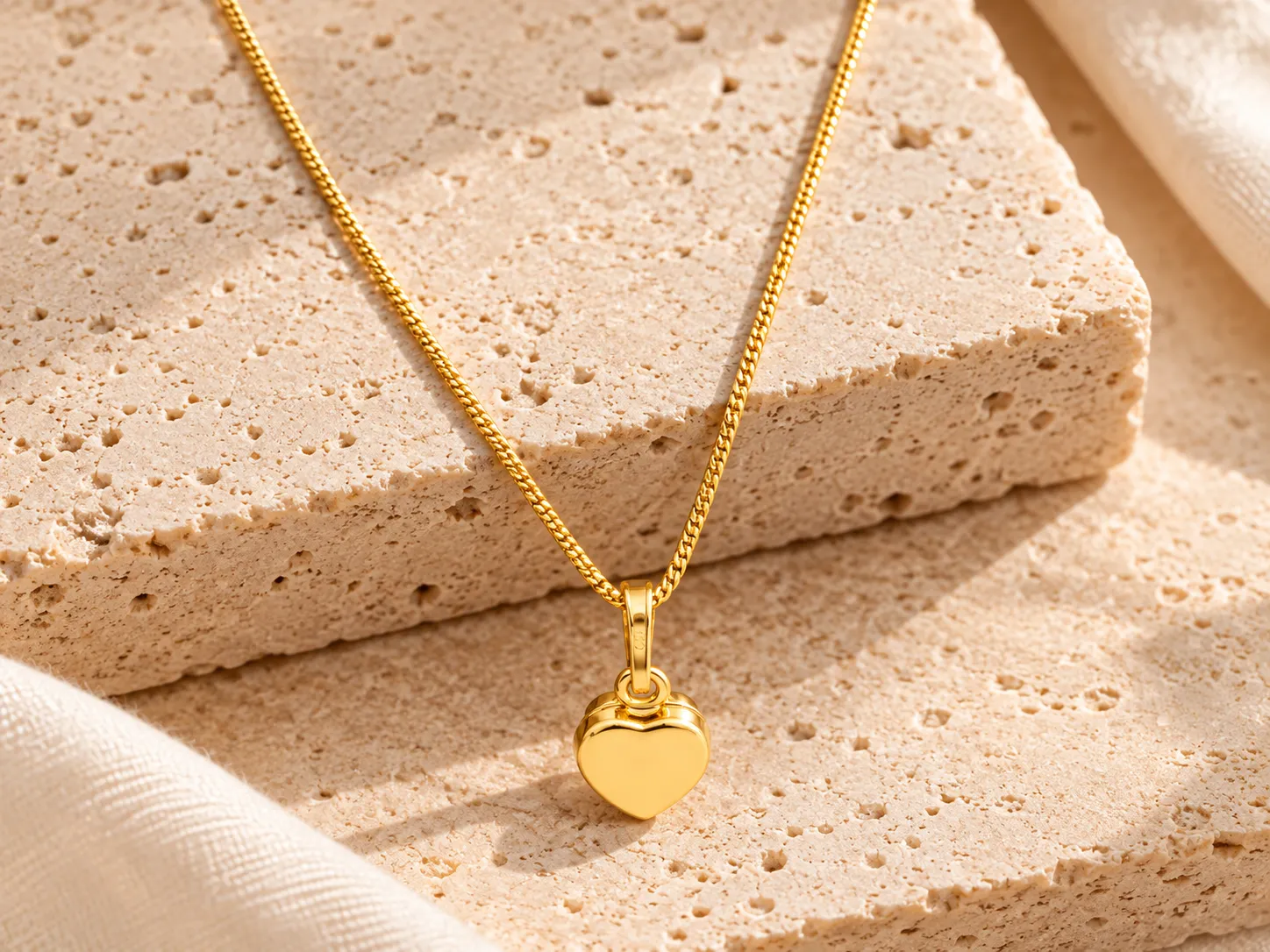 Gold necklace with a smooth puffed heart pendant on a curb chain, resting on a stone ledge