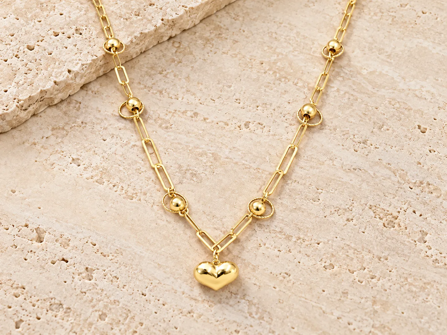 Gold paperclip-link necklace with small round beads and a puffed heart pendant, resting on a stone ledge