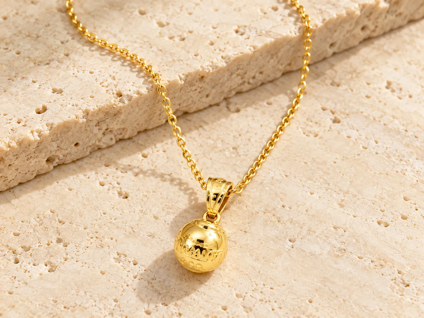 Gold necklace with a round, textured globe-shaped pendant on a fine chain, resting on a stone ledge