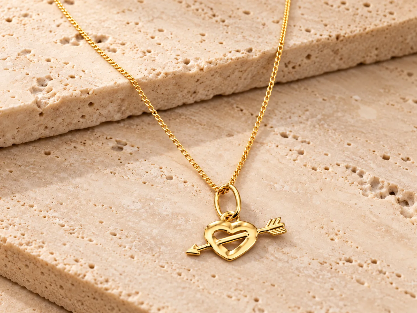 Gold necklace with a heart-and-arrow pendant on a fine curb chain, resting on a stone ledge