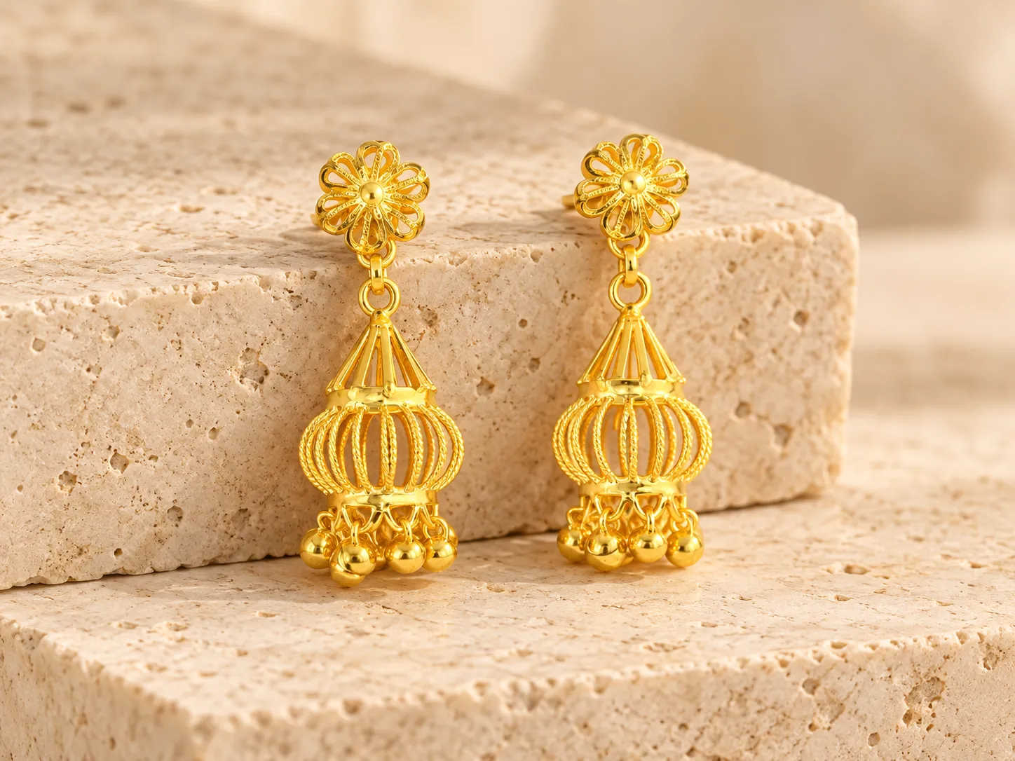A pair of gold lantern-style earrings with flower tops and hanging beads, resting on a stone ledge