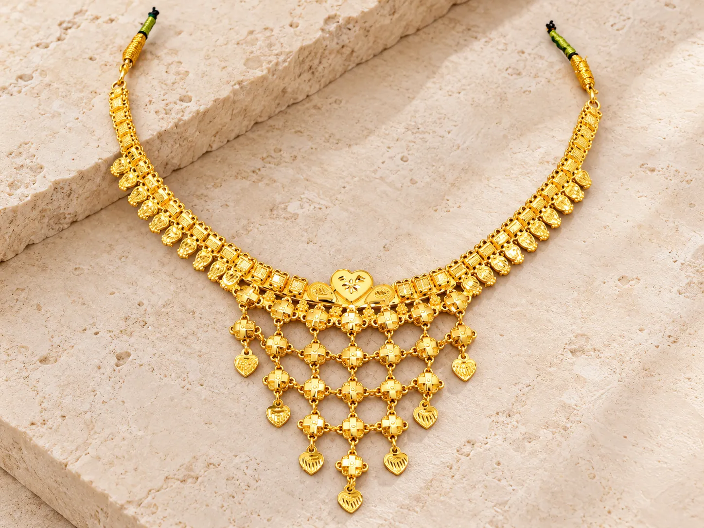 Traditional gold necklace with a heart-shaped centrepiece and rows of hanging heart-shaped drops, with green-and-gold cord ends, on a stone slab