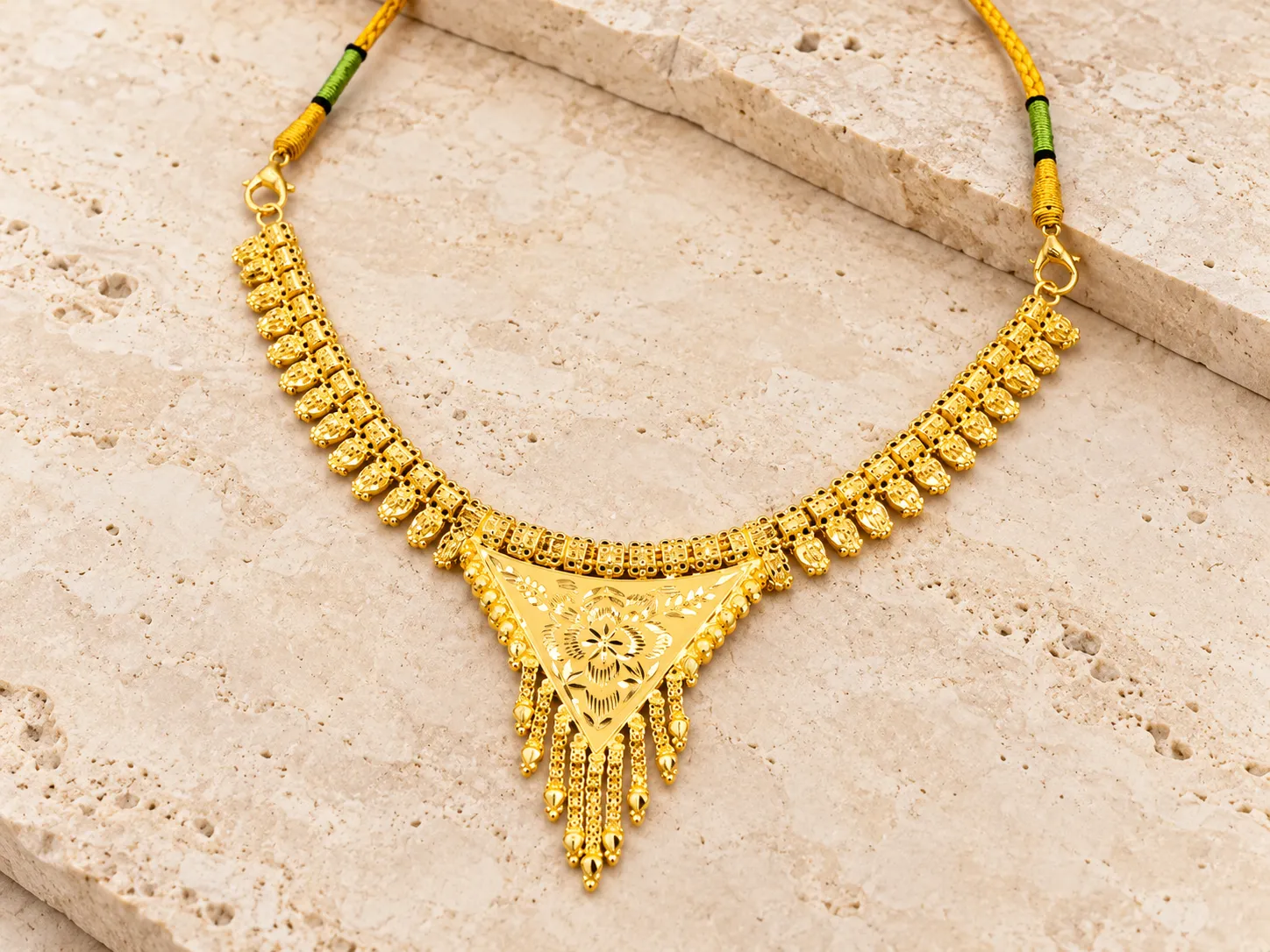 Traditional gold necklace with a floral centrepiece and hanging fringe, with green-and-gold cord ends, on a stone slab