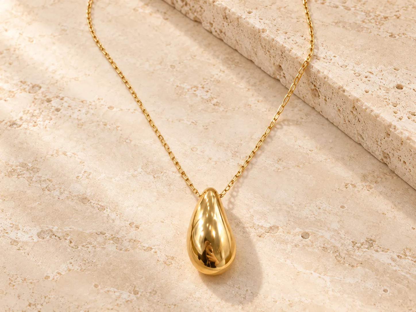 Gold necklace with a smooth, polished teardrop pendant on a fine chain, resting on a stone ledge