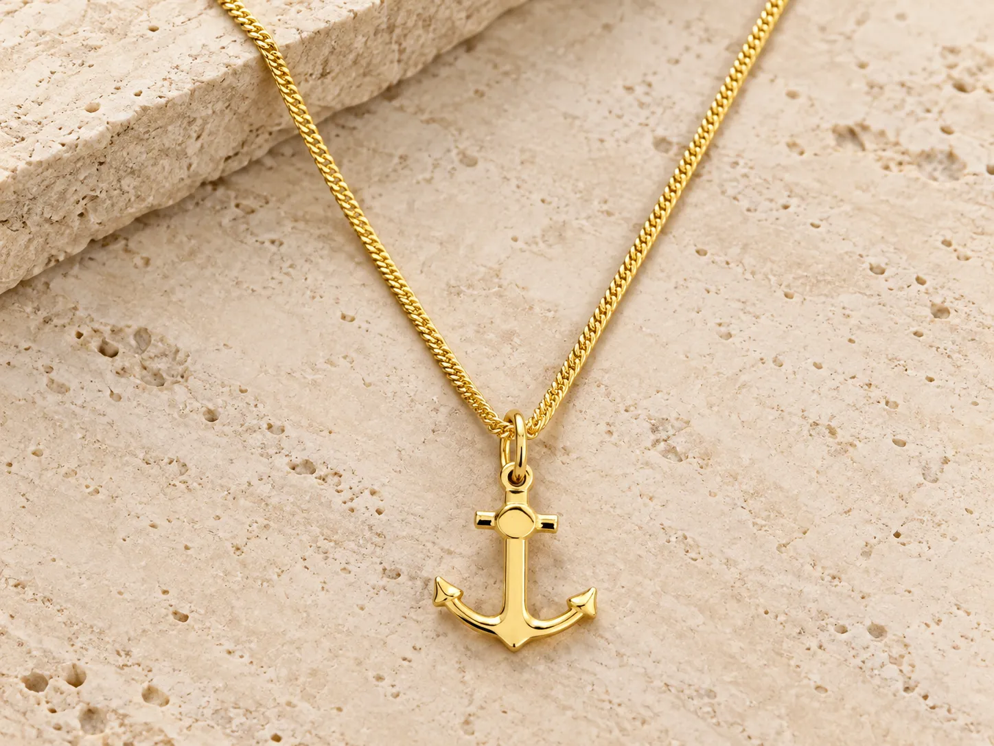 Gold necklace with an anchor-shaped pendant on a fine curb chain, resting on a stone ledge