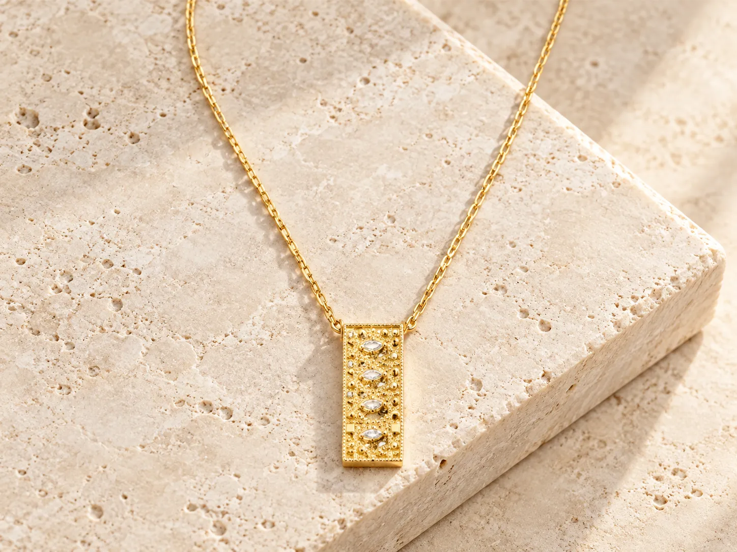 Gold necklace with a rectangular bar pendant with ornamental detailing, resting on a stone ledge