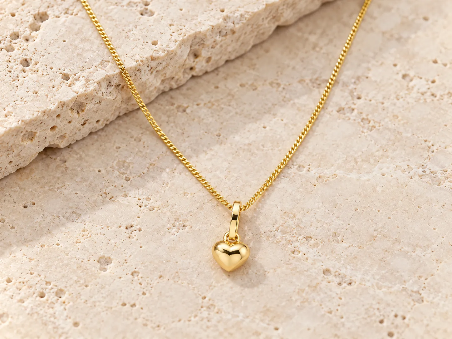 Gold necklace with a small heart pendant on a fine curb chain, resting on a stone ledge