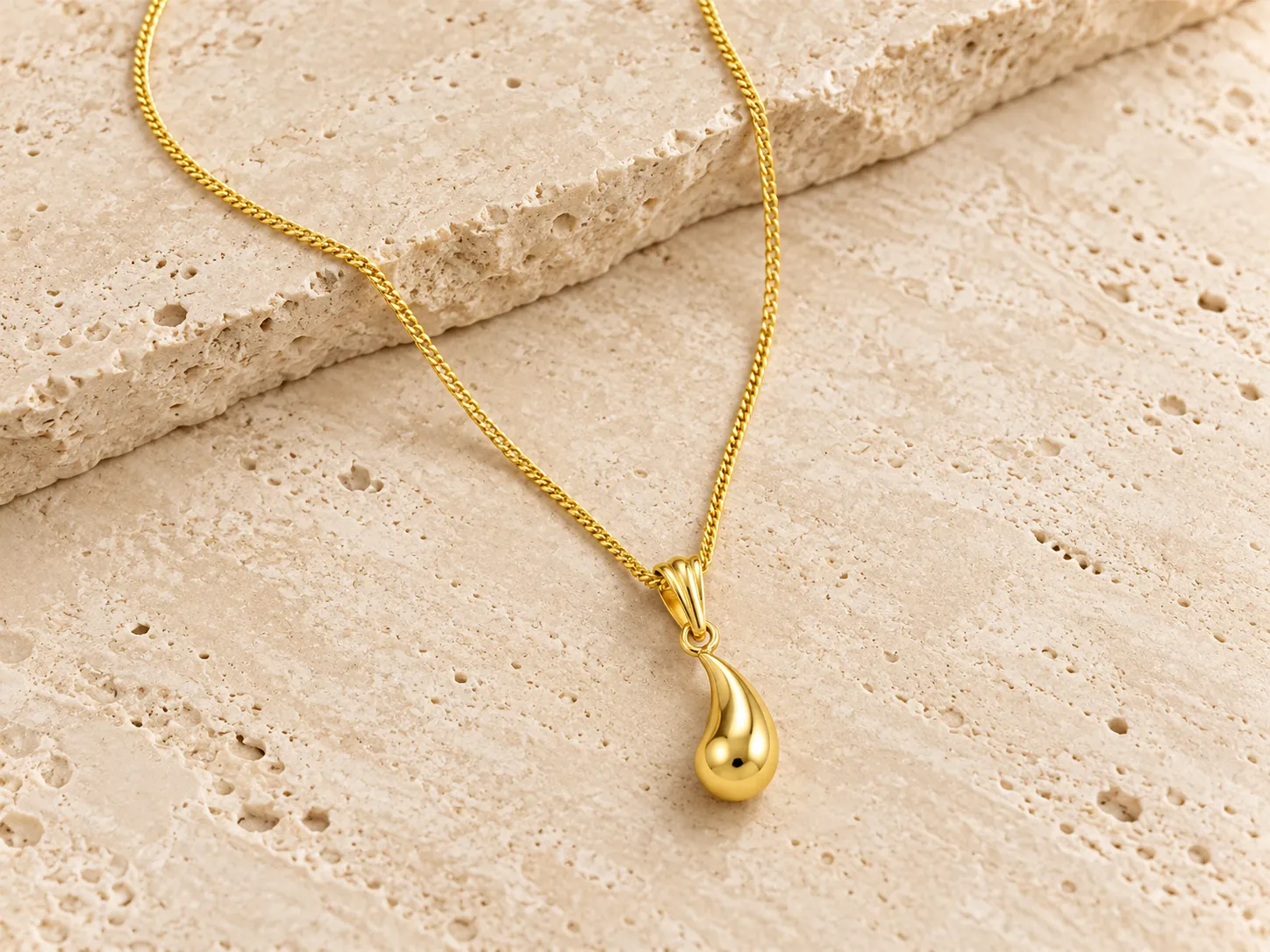Gold necklace with a smooth teardrop-shaped pendant on a fine curb chain, resting on a stone ledge