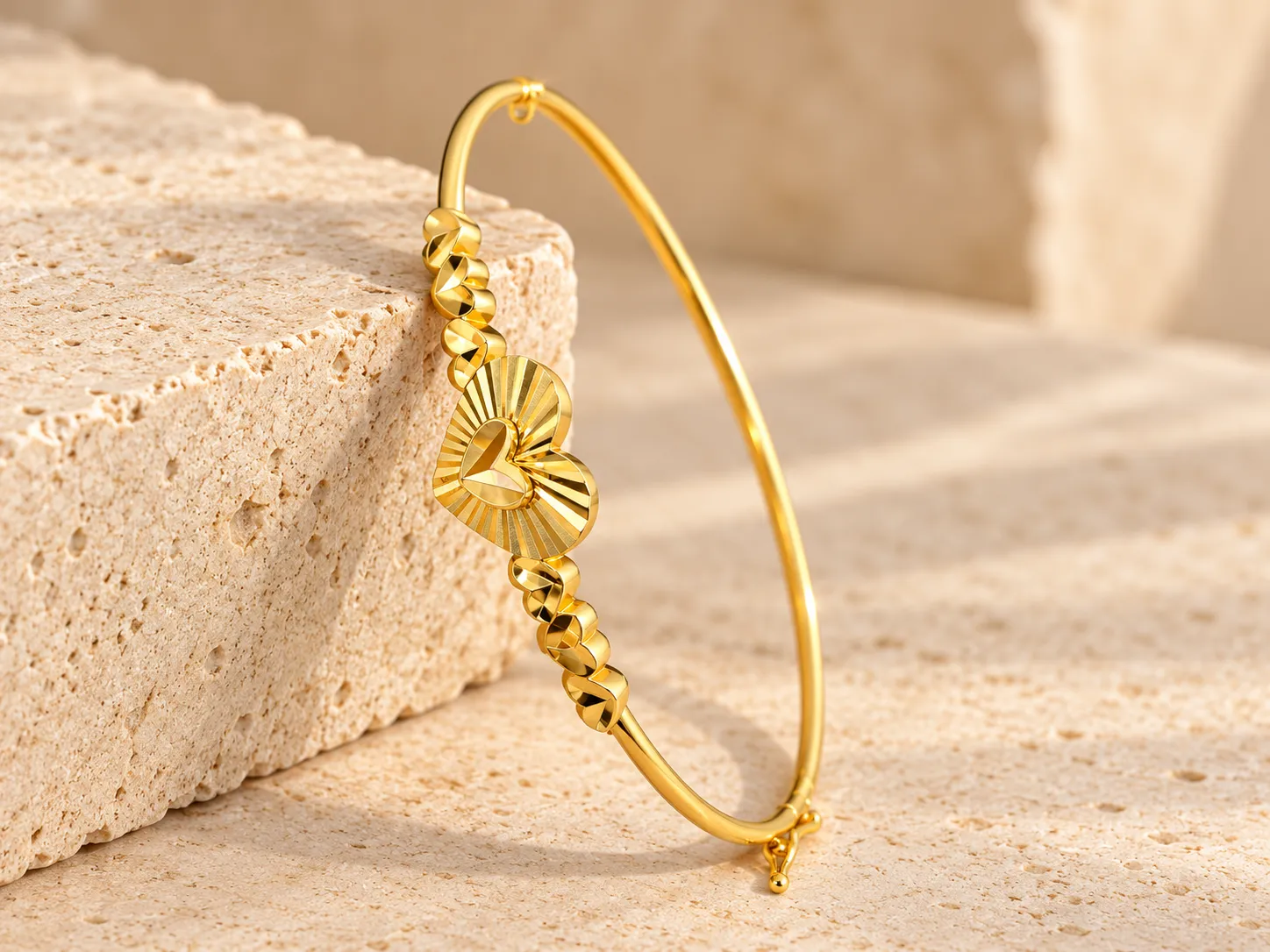 Slim gold bangle with a large heart centrepiece and twisted gold accents on each side, leaning on a stone block