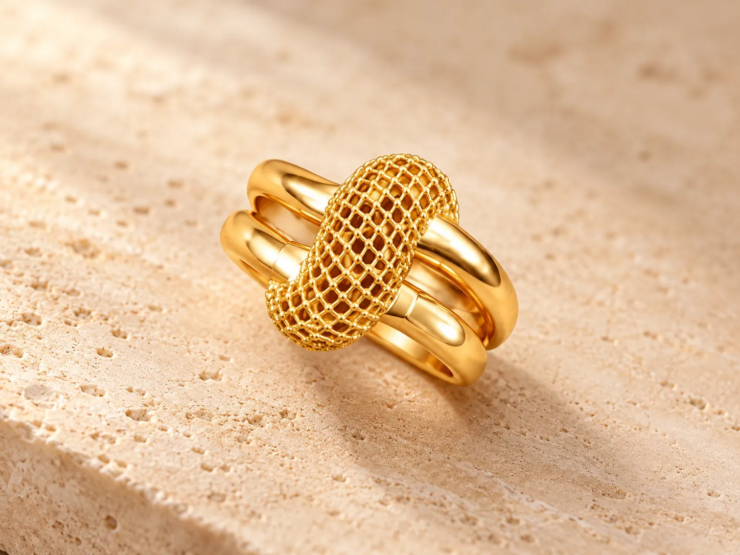 Gold ring with an oval mesh loop resting across polished gold bands, on a stone surface