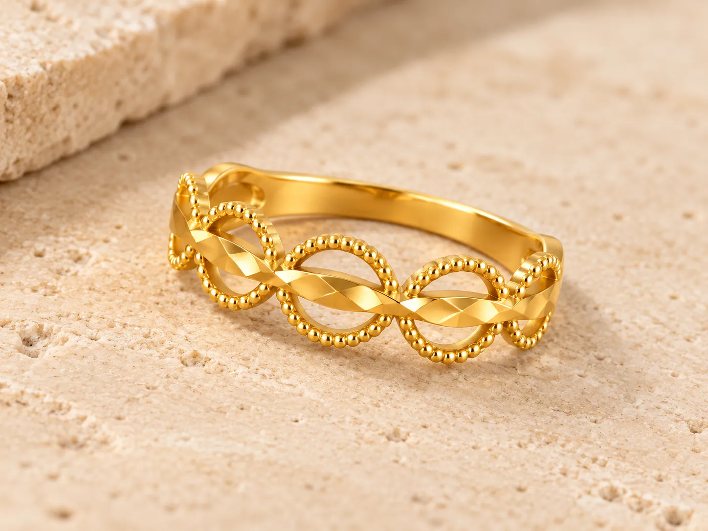 Slim gold ring of overlapping beaded loops crossed by a faceted band, on a stone surface