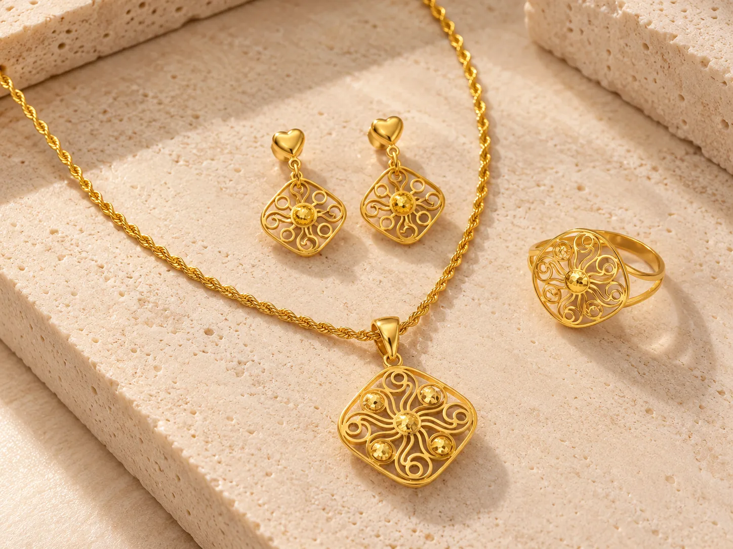 Gold rope chain necklace with a square scrollwork pendant beside heart-topped drop earrings and a round openwork ring, on a stone surface