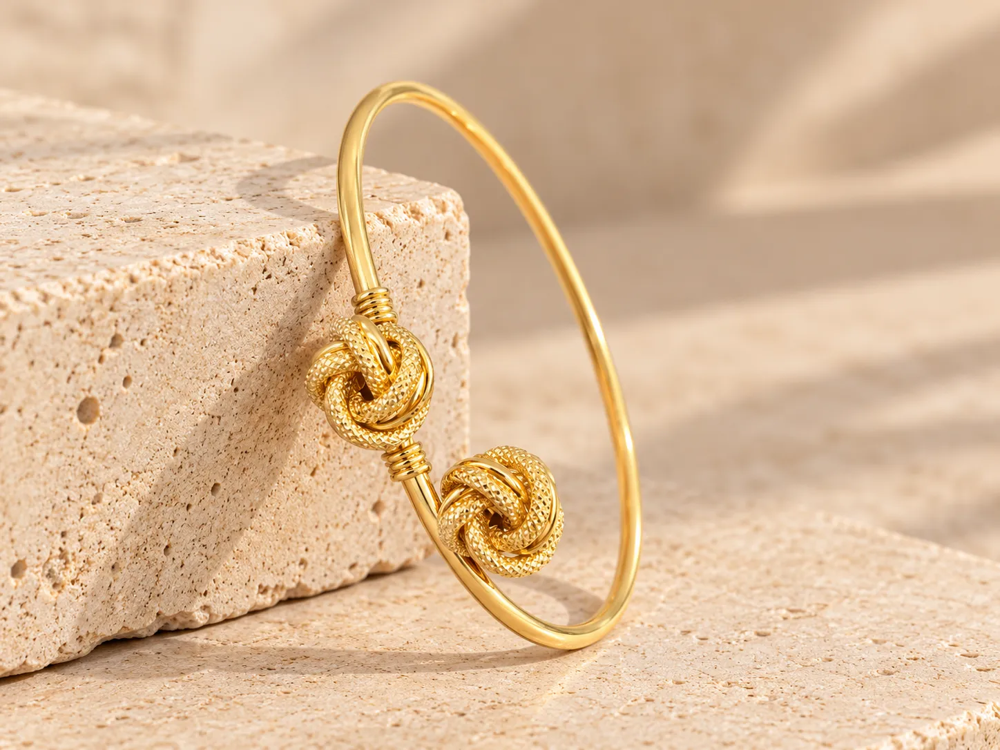 Slim open gold bangle ending in two textured gold knots, leaning on a stone block