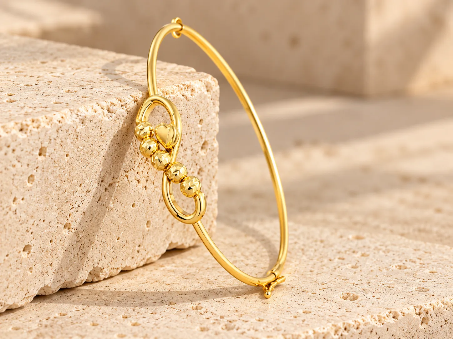 Slim gold bangle with a looped centre holding faceted beads and a small heart, leaning on a stone block