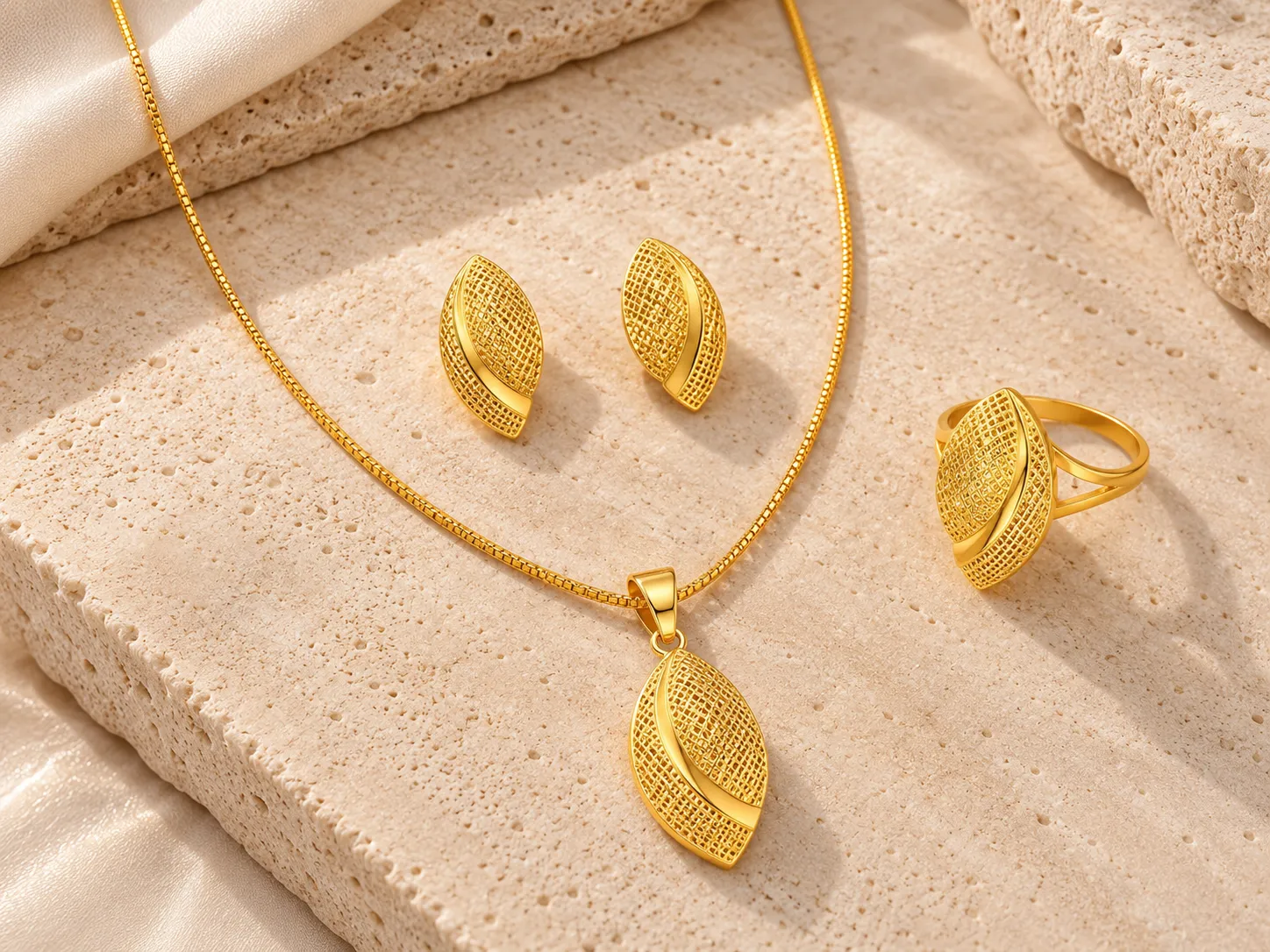 Gold necklace with a layered marquise-shaped mesh pendant beside matching earrings and a ring, on a stone surface