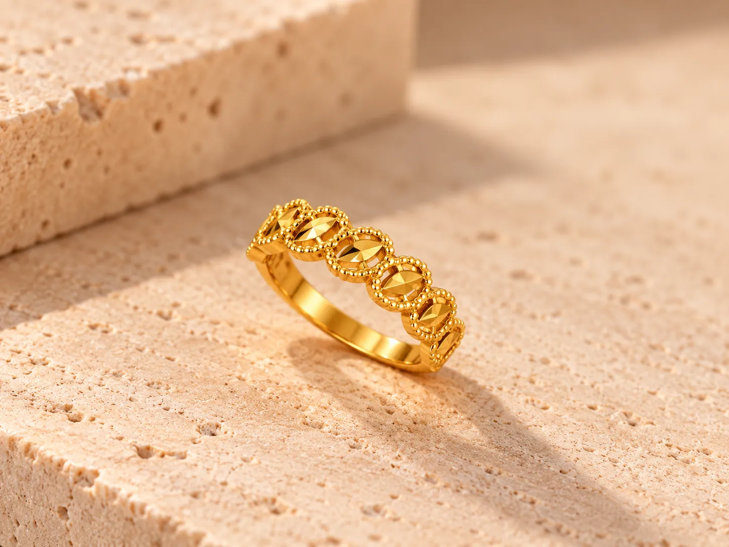 Gold band ring of linked beaded oval motifs with faceted gold centres, on a stone ledge