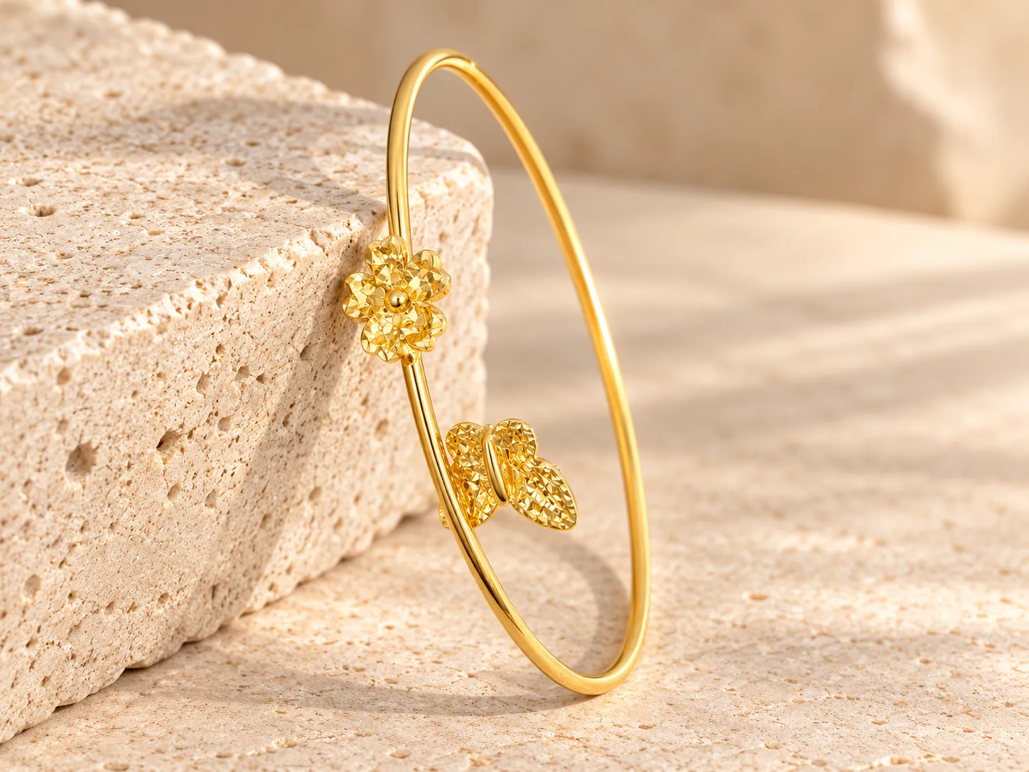 Slim open gold bangle with a textured flower and a pair of leaves at its ends, leaning on a stone block