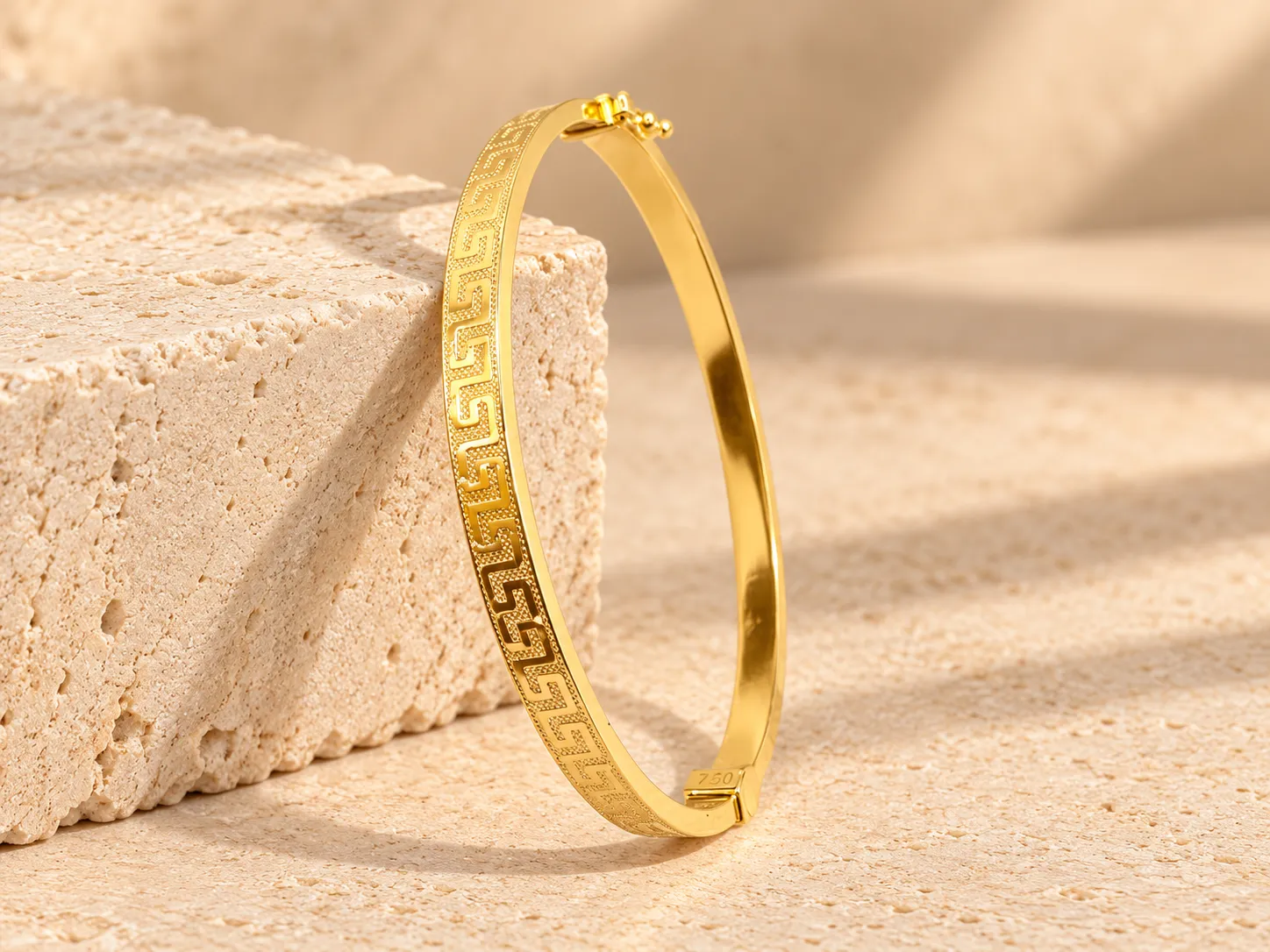 Gold hinged bangle with a Greek-key pattern on its outer face, leaning on a stone block