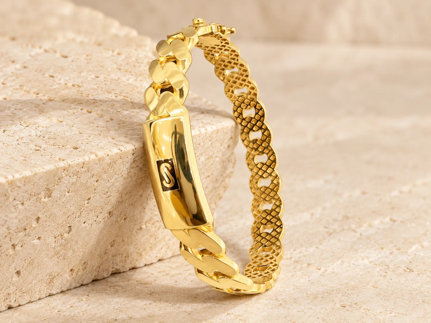 Gold bangle of broad curb-style links with a polished ID plate at the front, leaning on a stone block