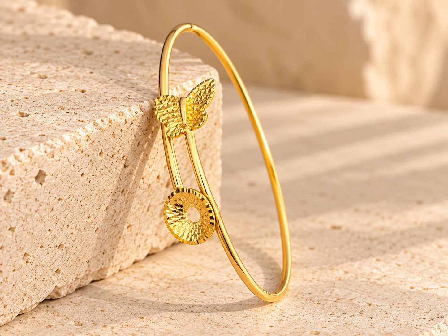 Slim open gold bangle with a textured butterfly and a textured round disc at its ends, leaning on a stone block