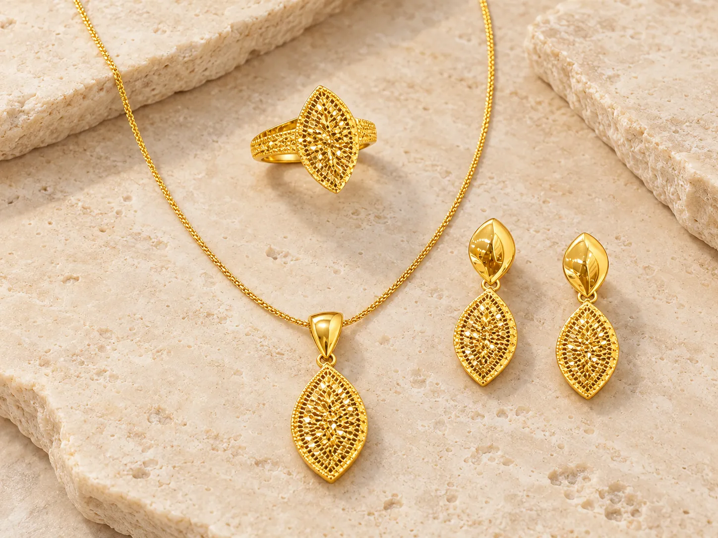Gold necklace with a marquise-shaped openwork pendant beside matching drop earrings and a ring, on a stone surface