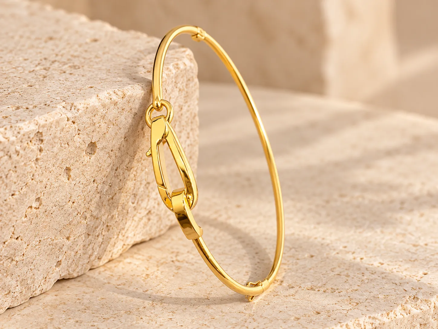 Slim gold bangle with a large elongated drop-shaped clasp link at the front, leaning on a stone block
