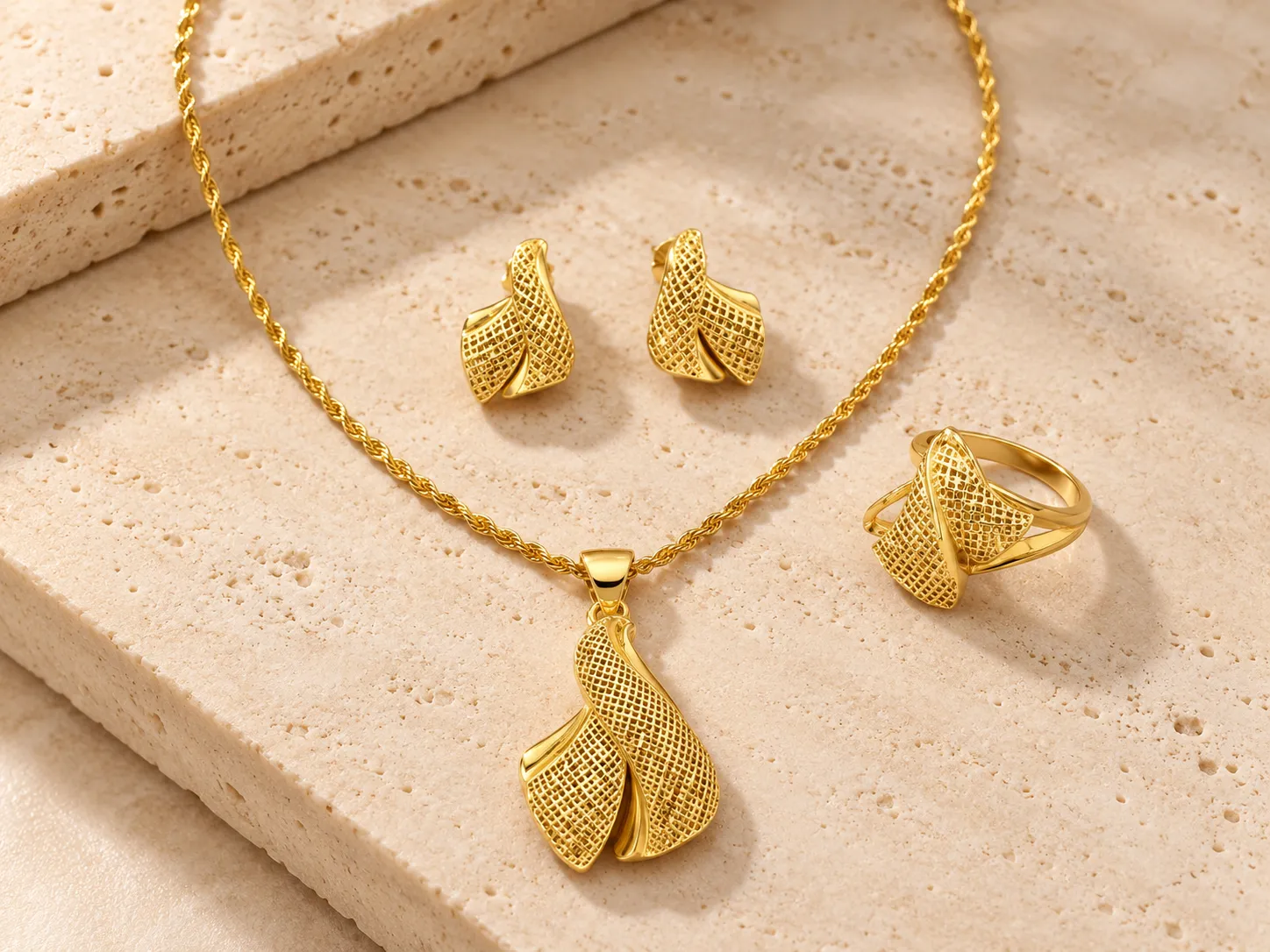 Gold rope chain necklace with a folded woven-mesh pendant beside matching earrings and a ring, on a stone surface