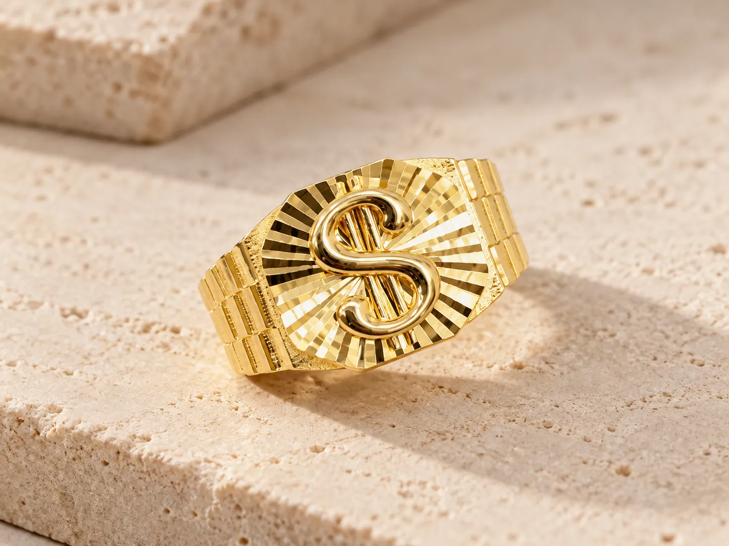 Gold ring with a polished S-shaped emblem on a face with radiating faceted lines and a link-style band, on a stone ledge