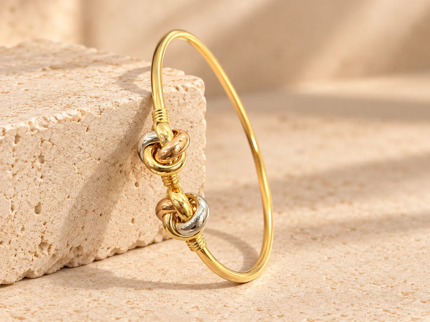 Slim gold bangle with two knots of yellow, white and rose gold rings, leaning on a stone block