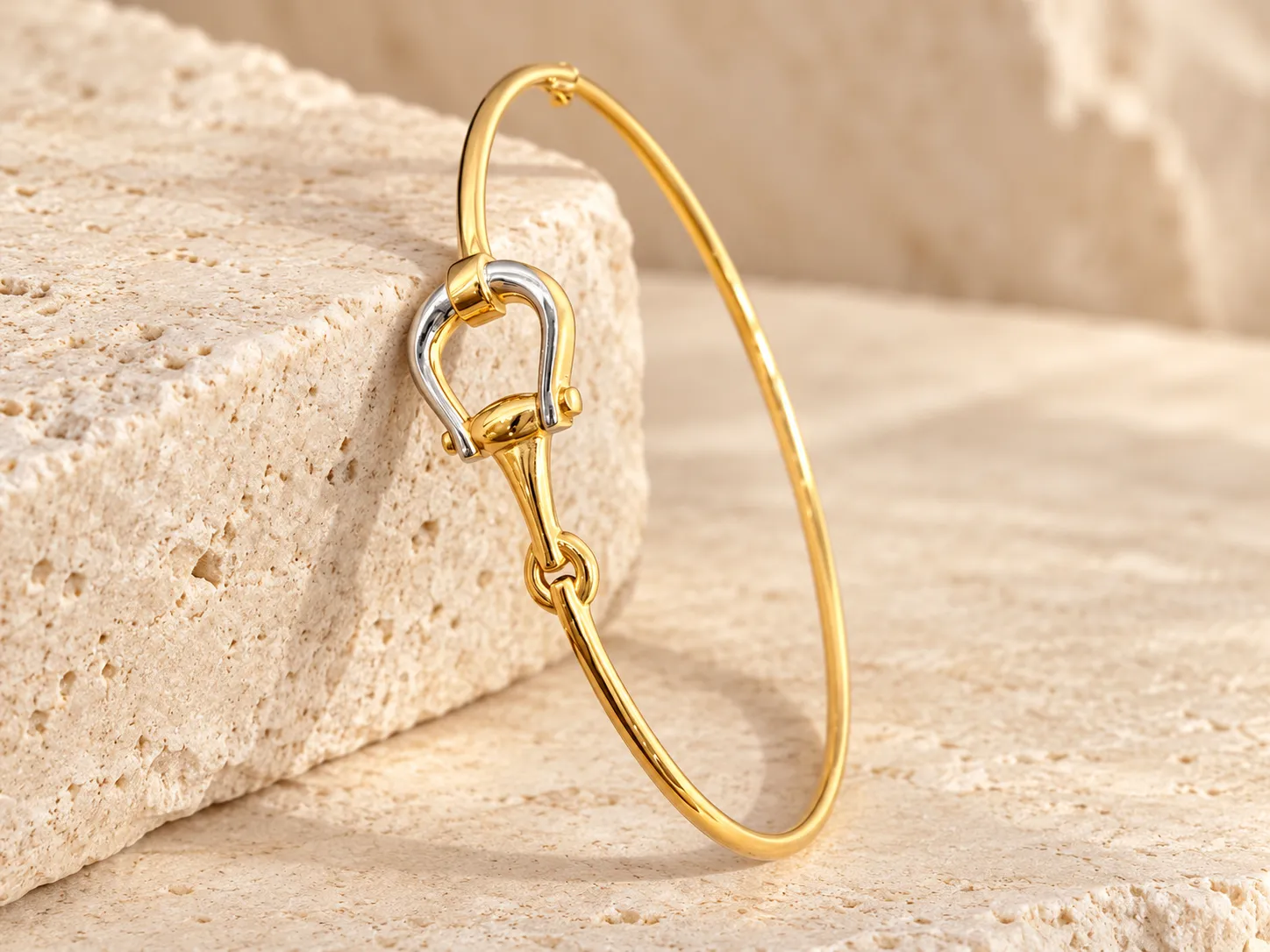 Slim gold bangle with a horseshoe-shaped white gold clasp and gold fitting at the front, leaning on a stone block