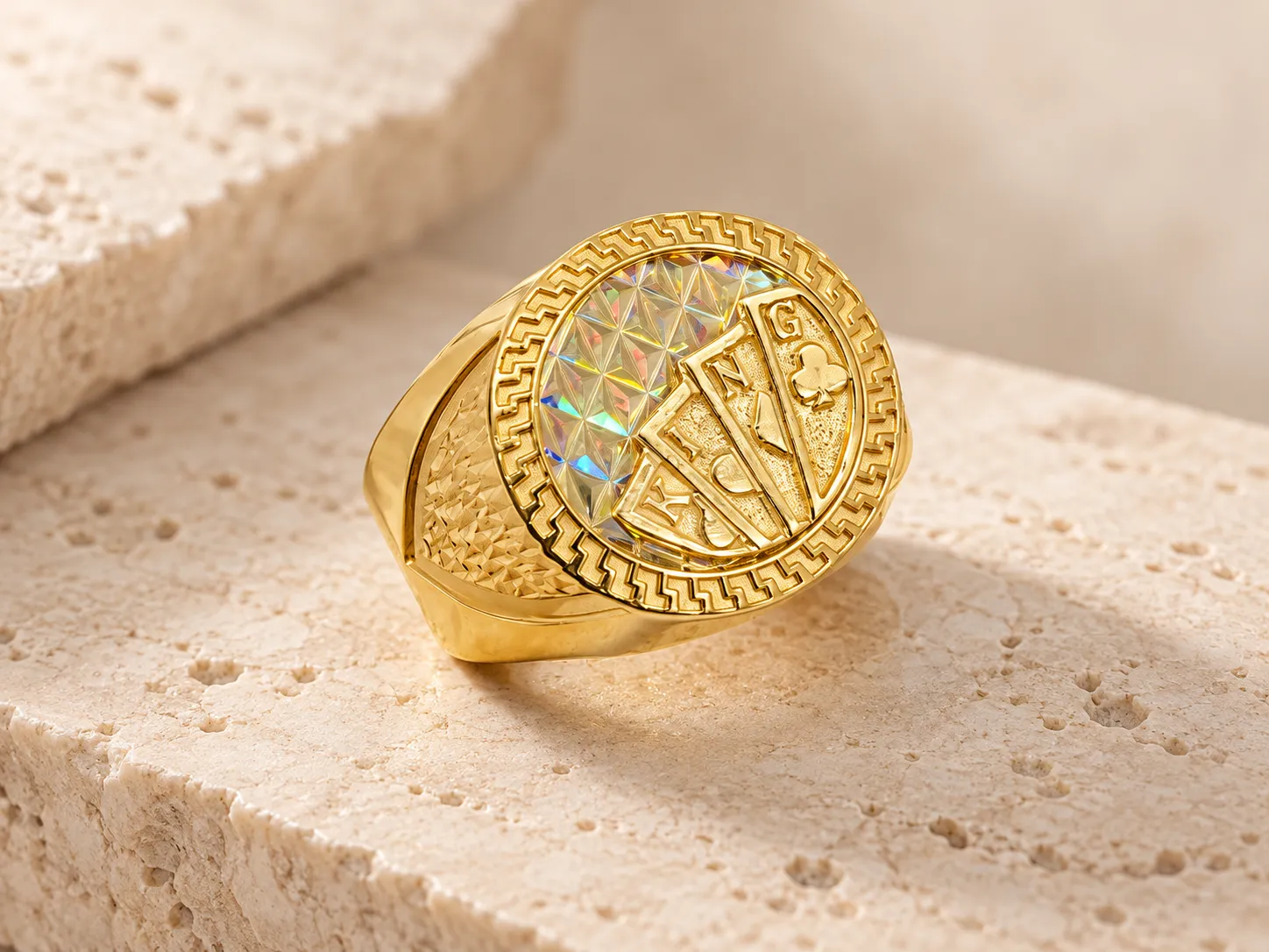Gold signet ring with a fan of raised playing cards on a prismatic oval face with a Greek-key border and textured shoulders, on a stone surface