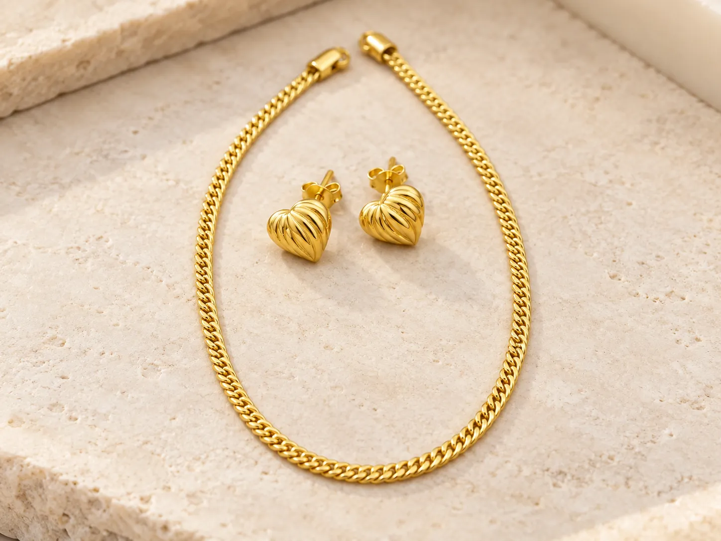 Gold curb-chain bracelet beside a pair of heart-shaped earrings on a warm stone surface