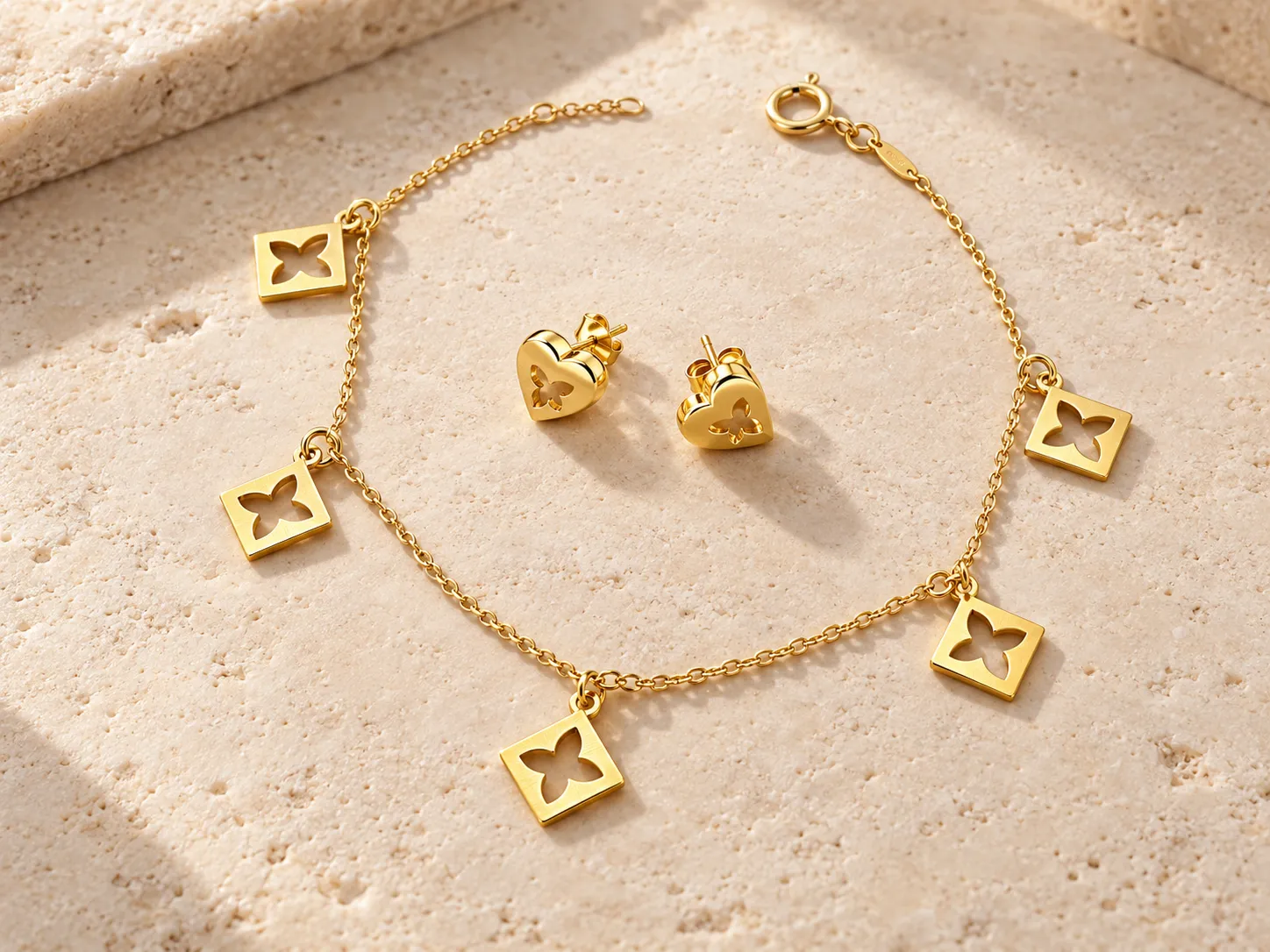 Gold charm bracelet with square cut-out charms beside a pair of heart-shaped earrings, on a warm stone surface