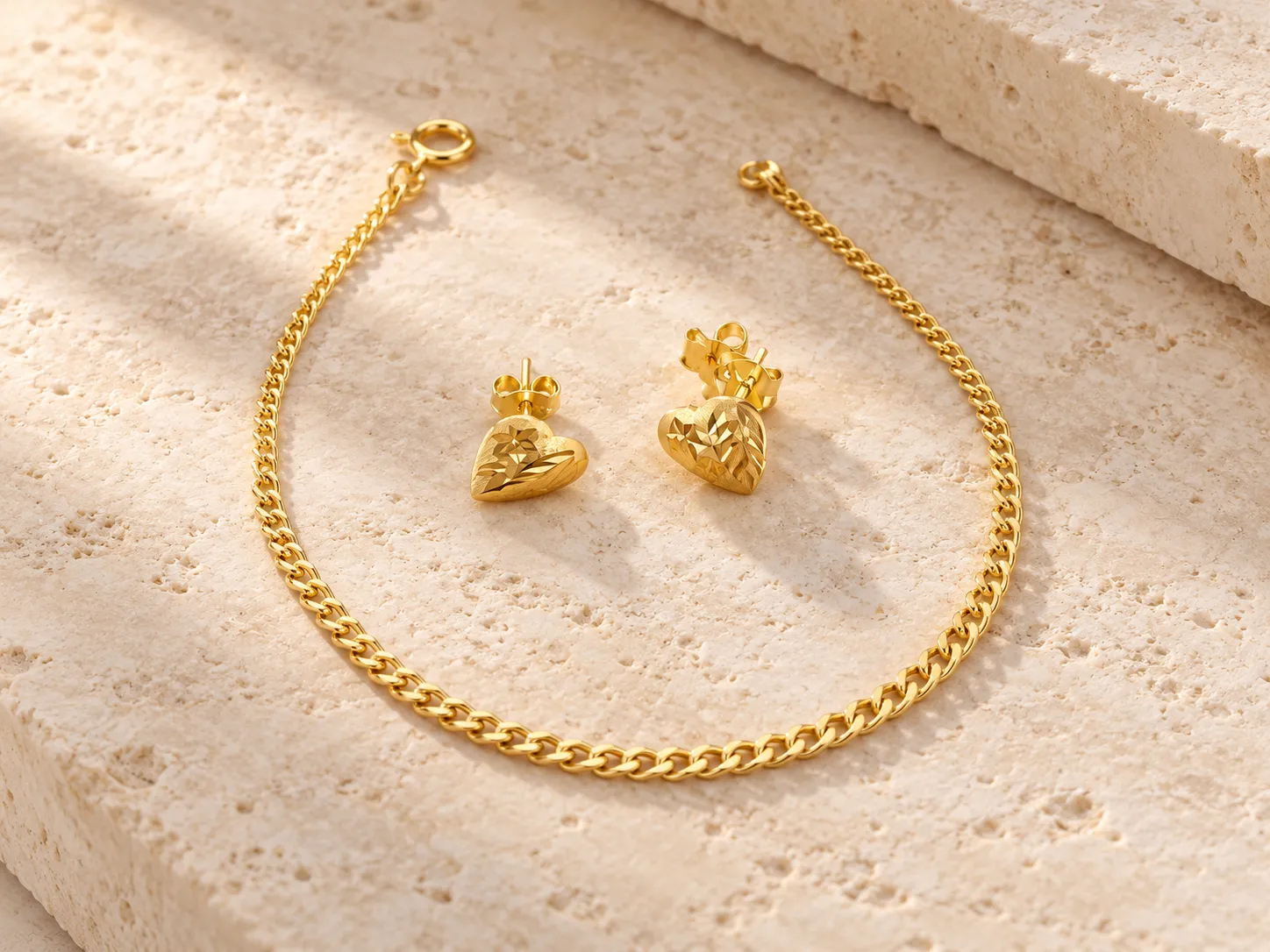 Gold curb-chain bracelet beside a pair of heart-shaped earrings on a warm stone surface