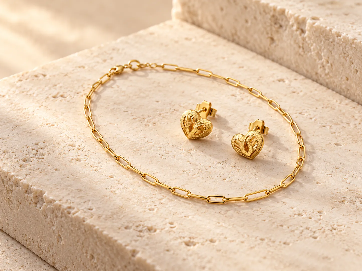 Gold paperclip-chain bracelet beside a pair of heart-shaped earrings on a warm stone surface