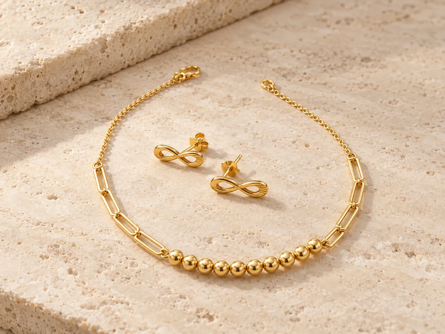 Gold bracelet with paperclip-chain links and a row of beads beside a pair of infinity-shaped earrings, on pale stone