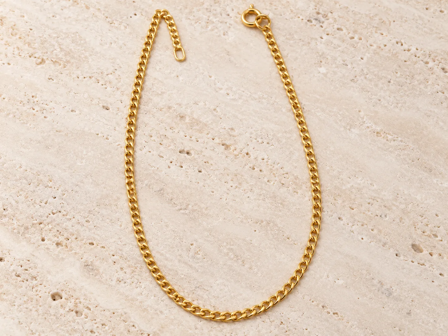 Gold curb-chain necklace laid in a loop on a warm stone surface