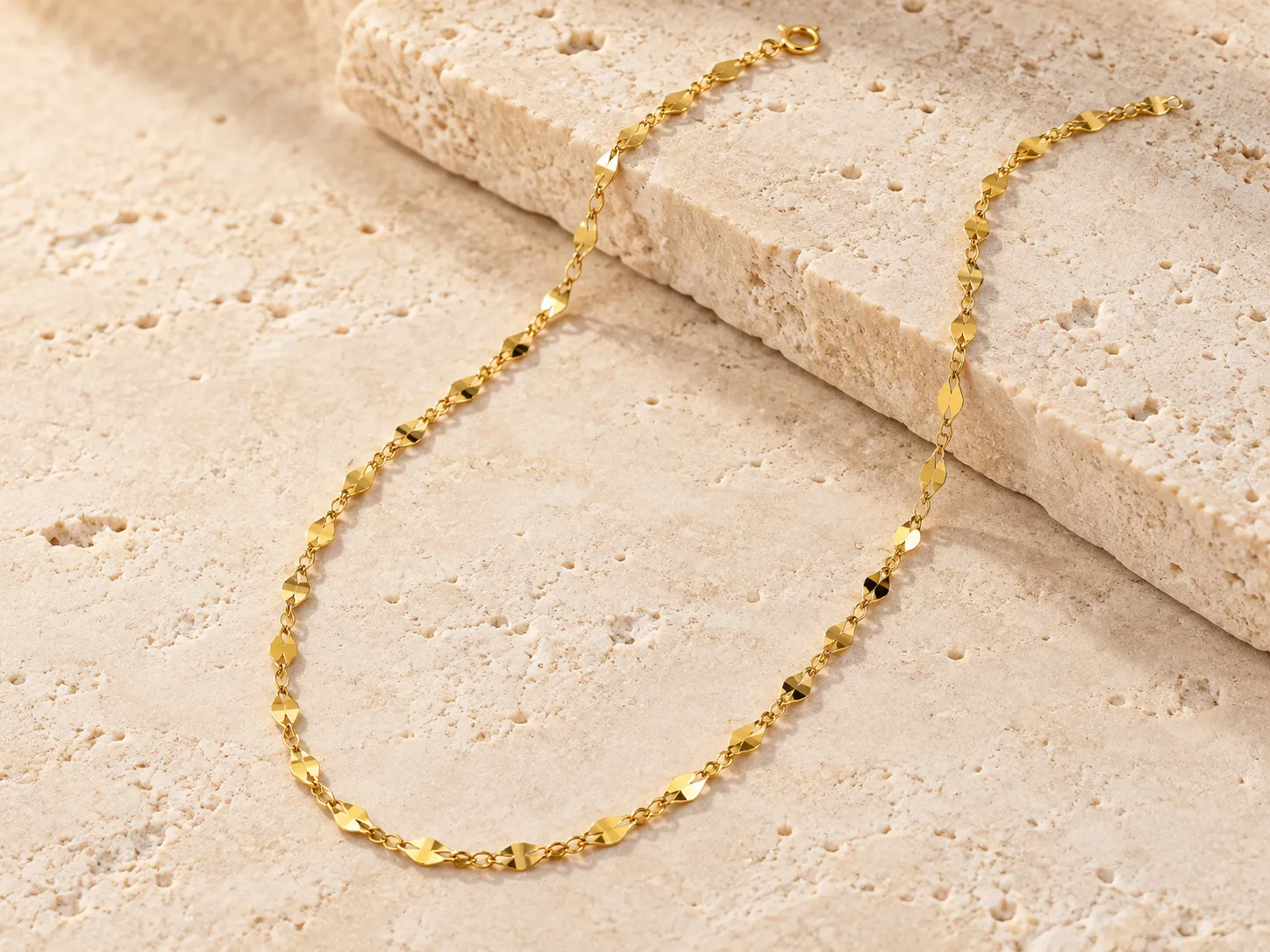 Gold chain necklace with small flat, faceted links on a warm stone surface